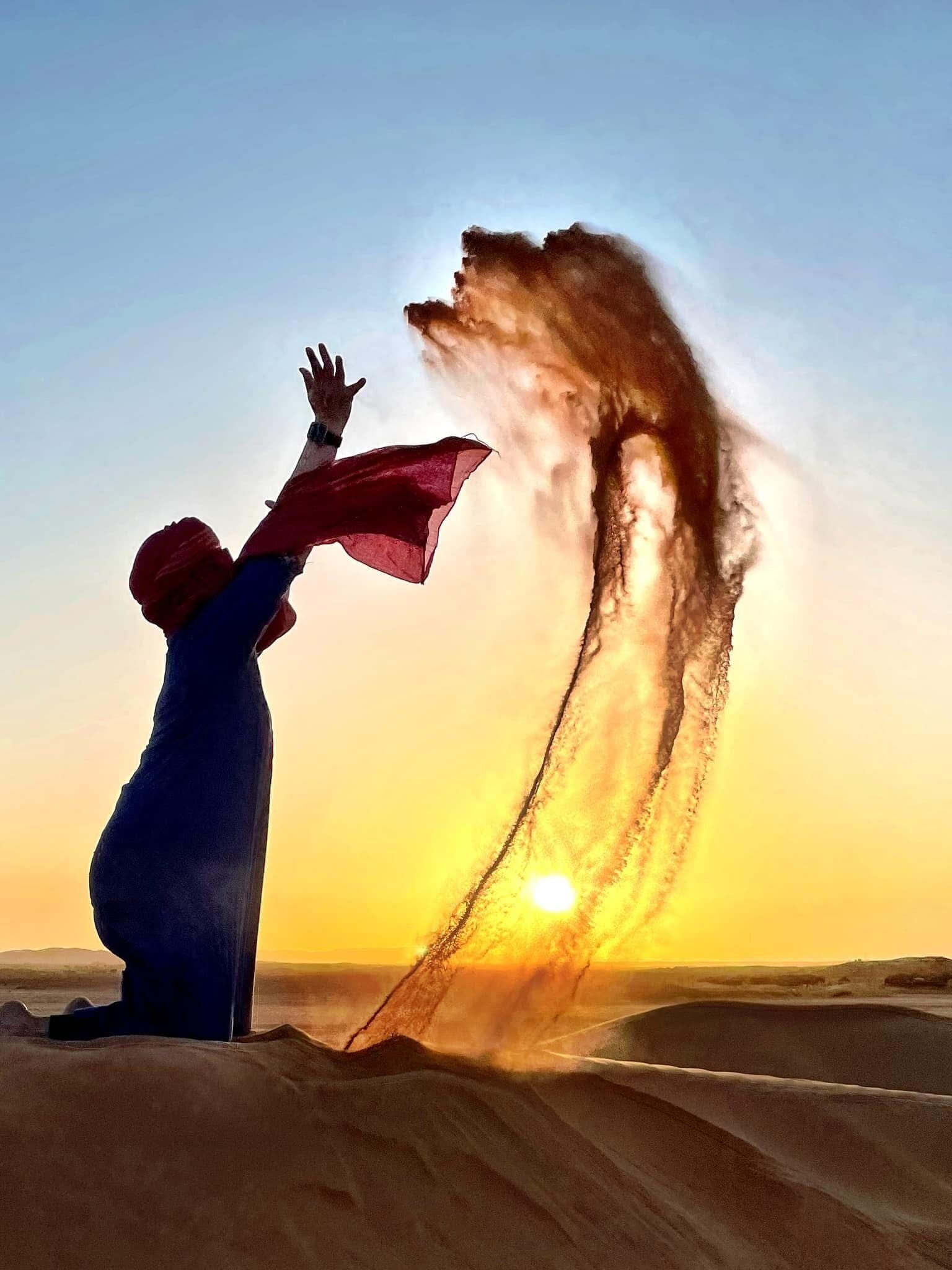 A person standing in a desert with sand dunes in the background and a clear sky.