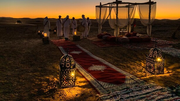 Cena no deserto com um grupo de pessoas vestindo trajes tradicionais, um toldo e um tapete vermelho que leva a uma área de estar com lanternas.