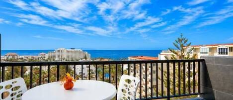 A balcony with a round table and chairs, a vase with flowers, and a view of the sea and buildings.