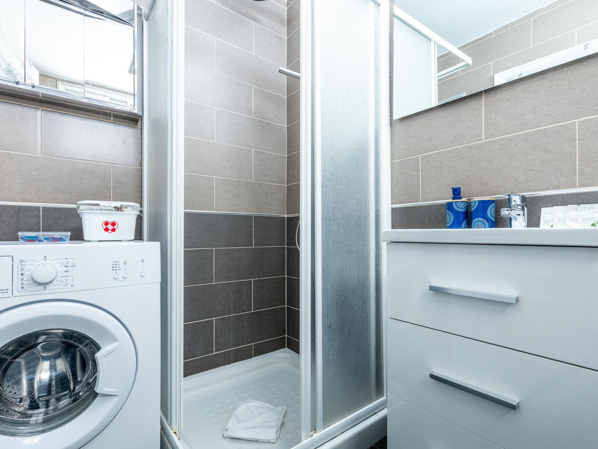A modern bathroom with a built-in washing machine, a shower enclosure, and a vanity with a mirror.