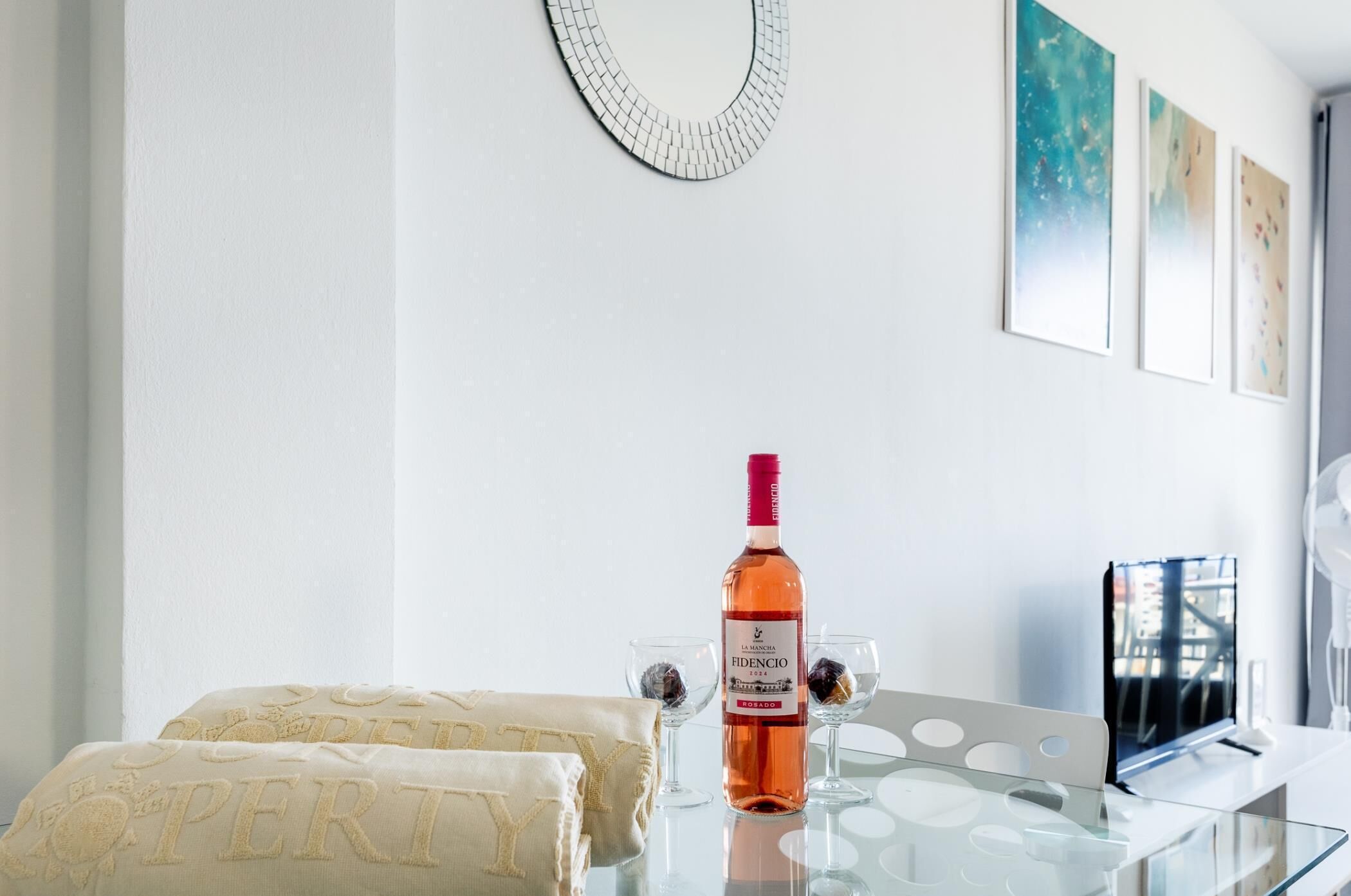 A modern hotel room with a glass table, a bottle of wine, two glasses, and a bed with pillows.