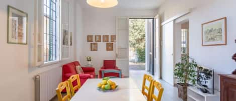 A dining area with a white table, yellow and blue chairs, and a fruit bowl. There are red chairs and a sofa in the background.