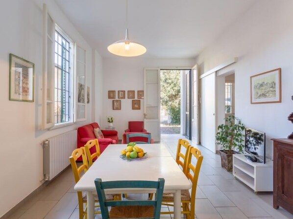 A dining area with a white table, yellow and blue chairs, and a fruit bowl. There are red chairs and a sofa in the background.