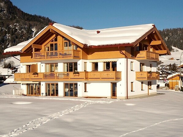 Un chalet à plusieurs étages, doté de balcons en bois et d’un toit enneigé, niché au cœur d’un paysage hivernal.