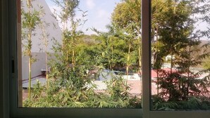 A window view of a garden with various plants and a red-roofed building in the background. - PALMAS DE CHAHUE 8 (1,100 SF) (Bahías de HUATULCO)