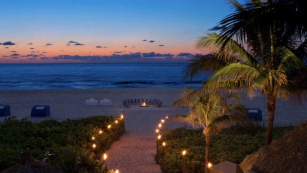 A beach pathway with a thatched-roof structure, palm trees, and a view of the ocean at dusk.
