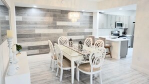 A modern kitchen with white cabinets, a central island, and a dining area with a glass table and white chairs. - : Sandy Feet Retreat: Walk to the Beach! : (Jupiter)
