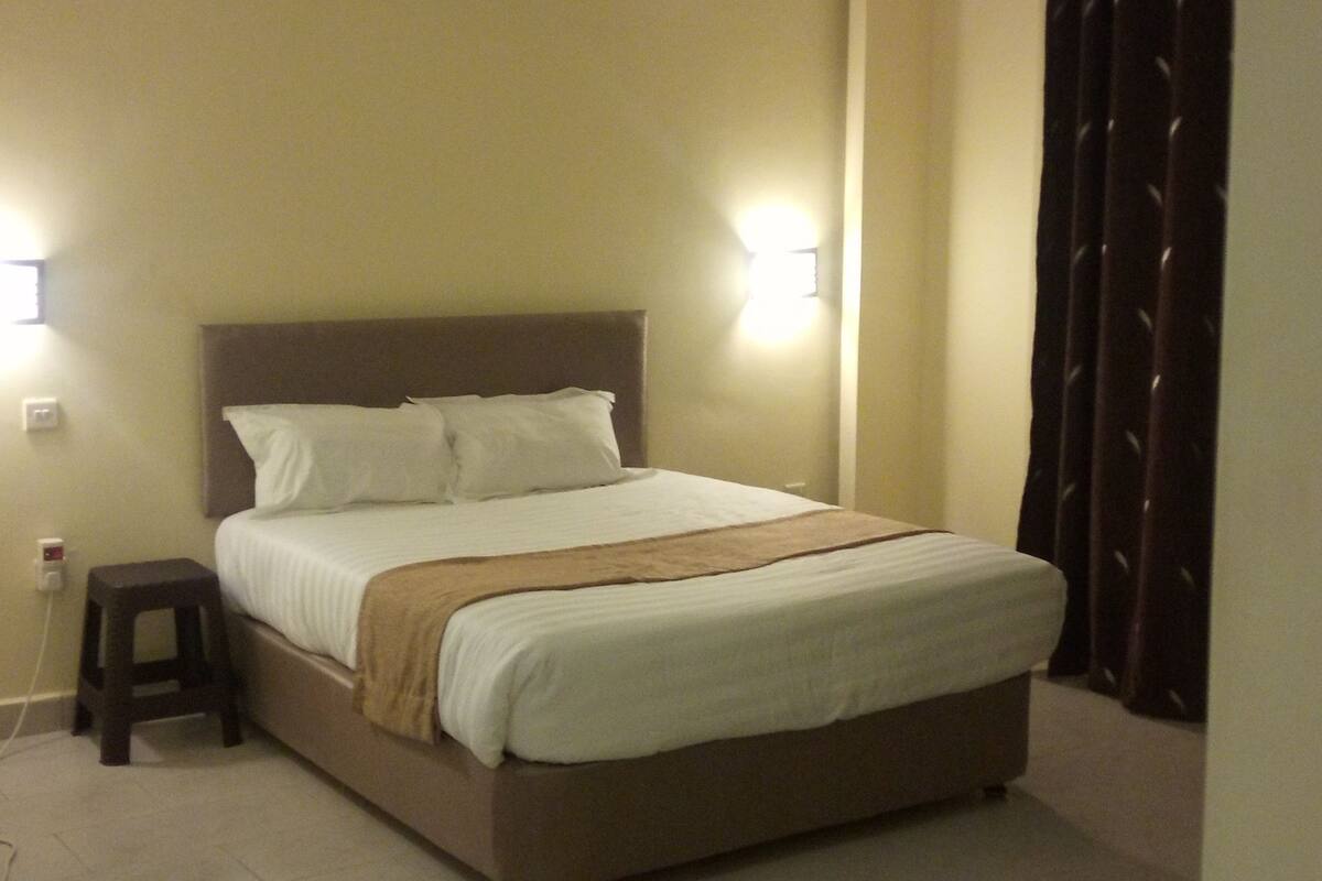 A hotel room with a large bed, a nightstand, a chair, and a wall-mounted light.