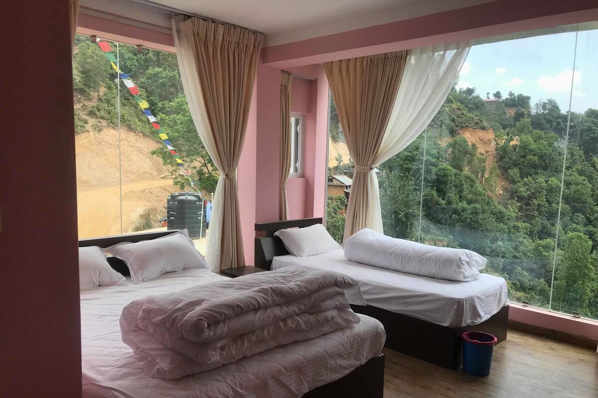 A hotel room with two beds, a large window with curtains, a view of a hillside, and a water tank outside.