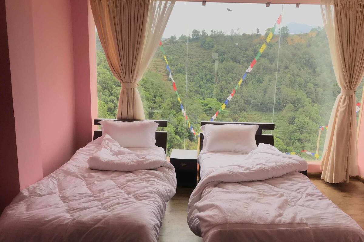 A room with two beds, a window with curtains, and a view of a green landscape.