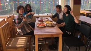 Un grupo de personas sentadas en una mesa de madera con varios platos, en una habitación con ventanas amplias y vistas a un paisaje montañoso.