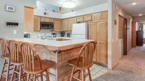 A modern kitchen with wooden cabinets, a central island with bar stools, a refrigerator, and a microwave. - Condo with Mountain Views! Shared Hot Tub! Shuttle Access! (Mammoth Lakes)