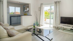 Una sala de estar moderna con un sofá, una mesa de centro de vidrio, un televisor y una cocina con armarios y una ventana.