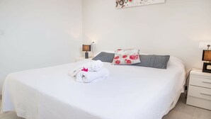 Una cama bien hecha con una colcha blanca, una toalla doblada y un almohadón con estampado rojo y blanco.