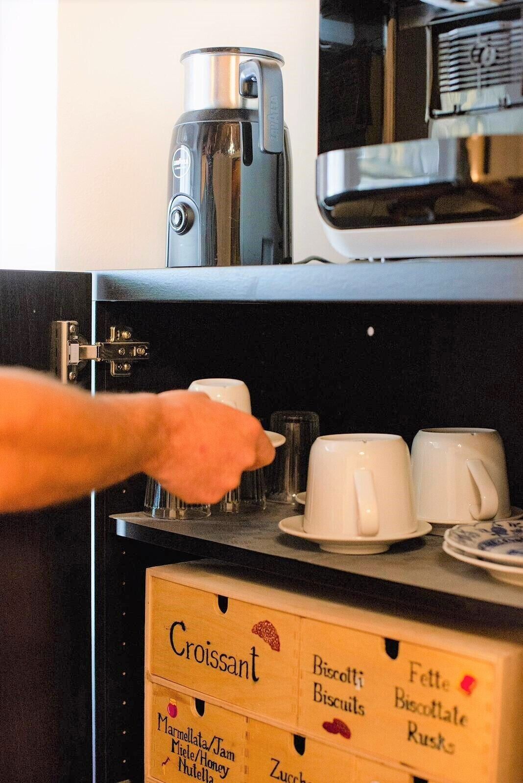 A kitchen area with a coffee machine, a hand reaching for a cup, and boxes of croissants and biscuits.