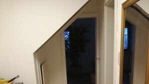 Un pasillo estrecho con piso de madera, paredes blancas y una habitación oscura al final.