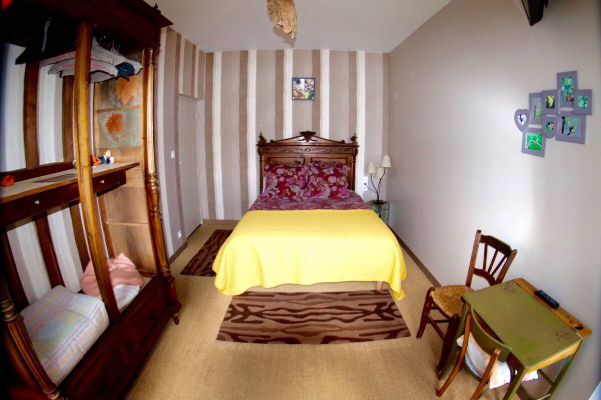 Ein Schlafzimmer mit einem Bett, einem Holzstuhl, einem Schreibtisch, einer Lampe und einer Wand mit gerahmten Bildern.