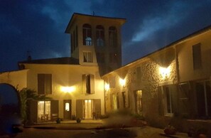 A building with a tower and multiple windows, some with shutters, and a well-lit entrance. - Chateau Trillon (Sauternes)