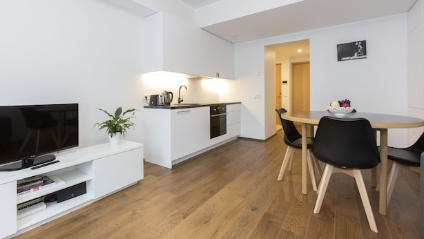 Eine moderne Küche mit weißen Schränken, einer schwarzen Arbeitsplatte und einem eingebauten Ofen. Es gibt einen Essbereich mit einem Holztisch und schwarzen Stühlen. Ein Flachbildfernseher steht auf einer weißen Unterhaltungsecke.