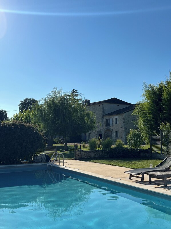 Une piscine avec des chaises longues, un bâtiment en pierre et un ciel d’un bleu limpide.