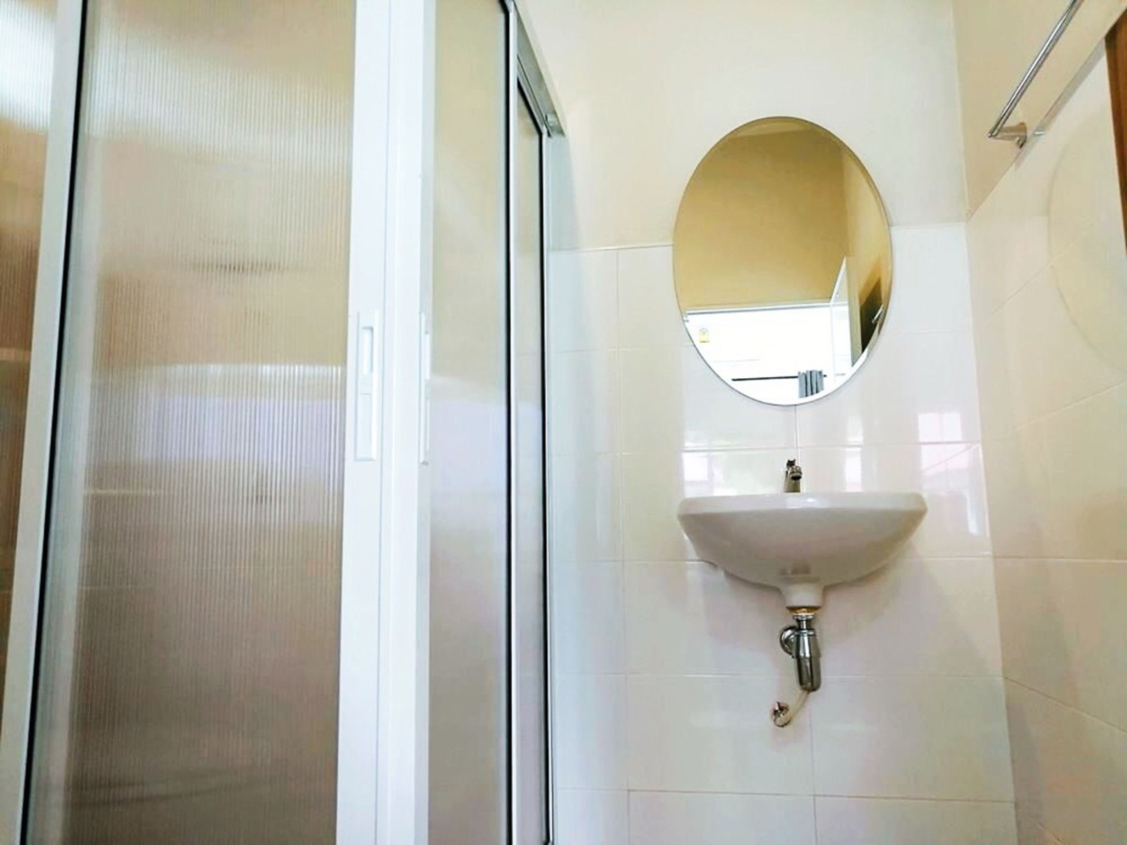 Un baño con mampara de vidrio, un lavamanos blanco y un espejo redondo encima, además de paredes con azulejos blancos.