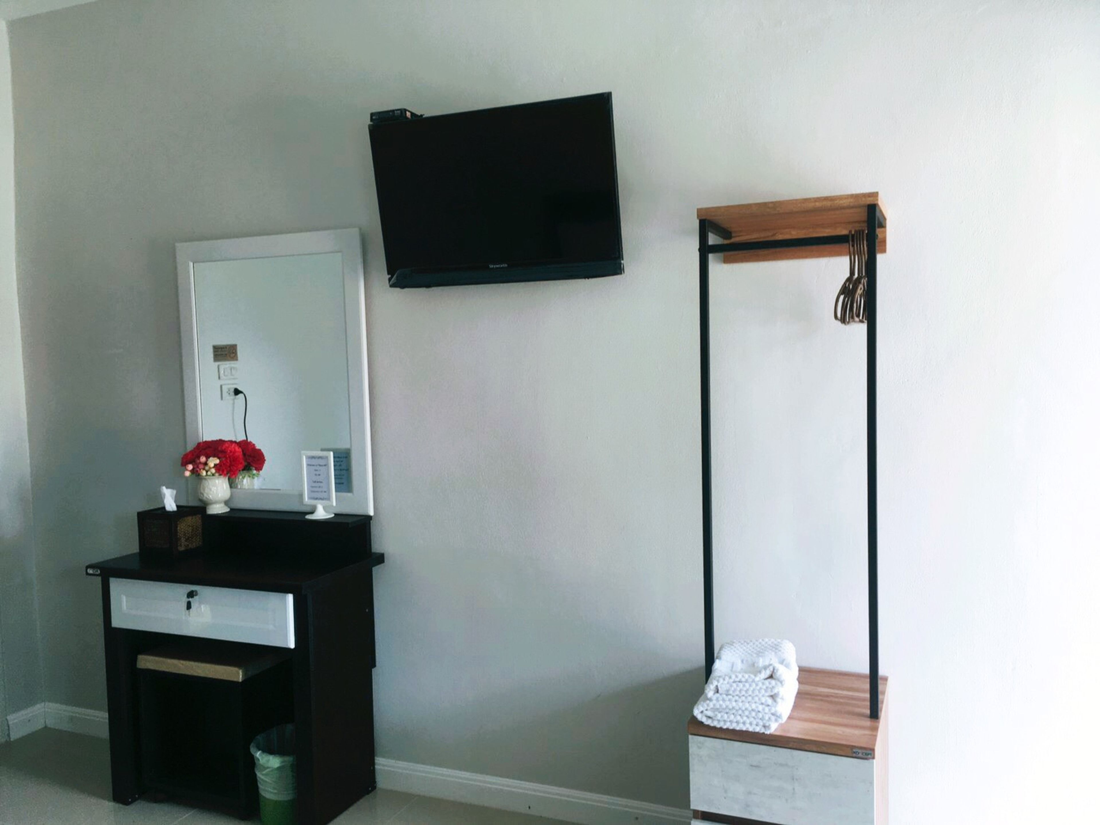 A room with a dark wooden dresser, a white vase with red flowers, a flat-screen TV mounted on the wall, and a wooden bench with a towel on it.