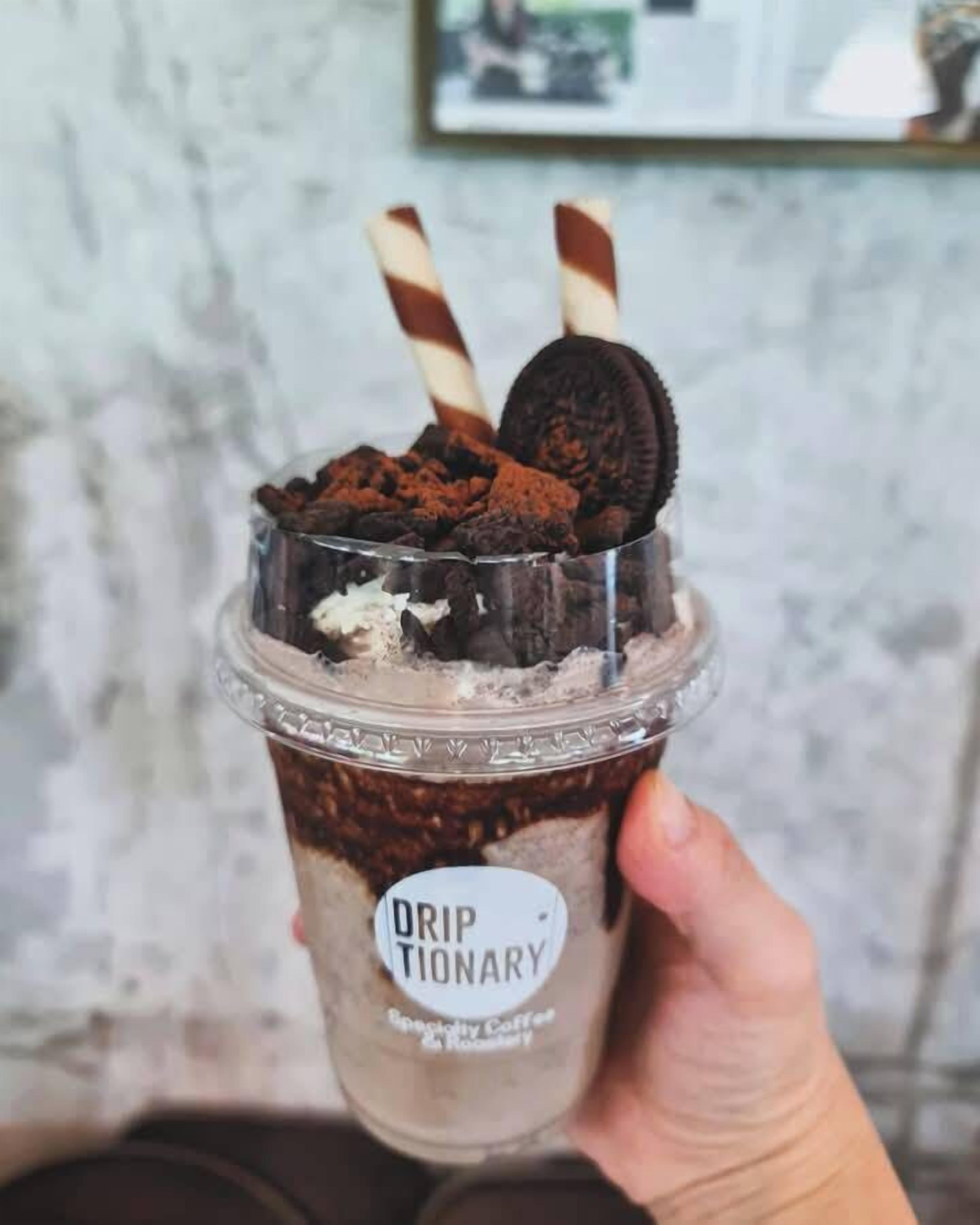 A hand holding a layered drink with a label that reads "Drip Tionary Specialty Coffee & Rosemary."
