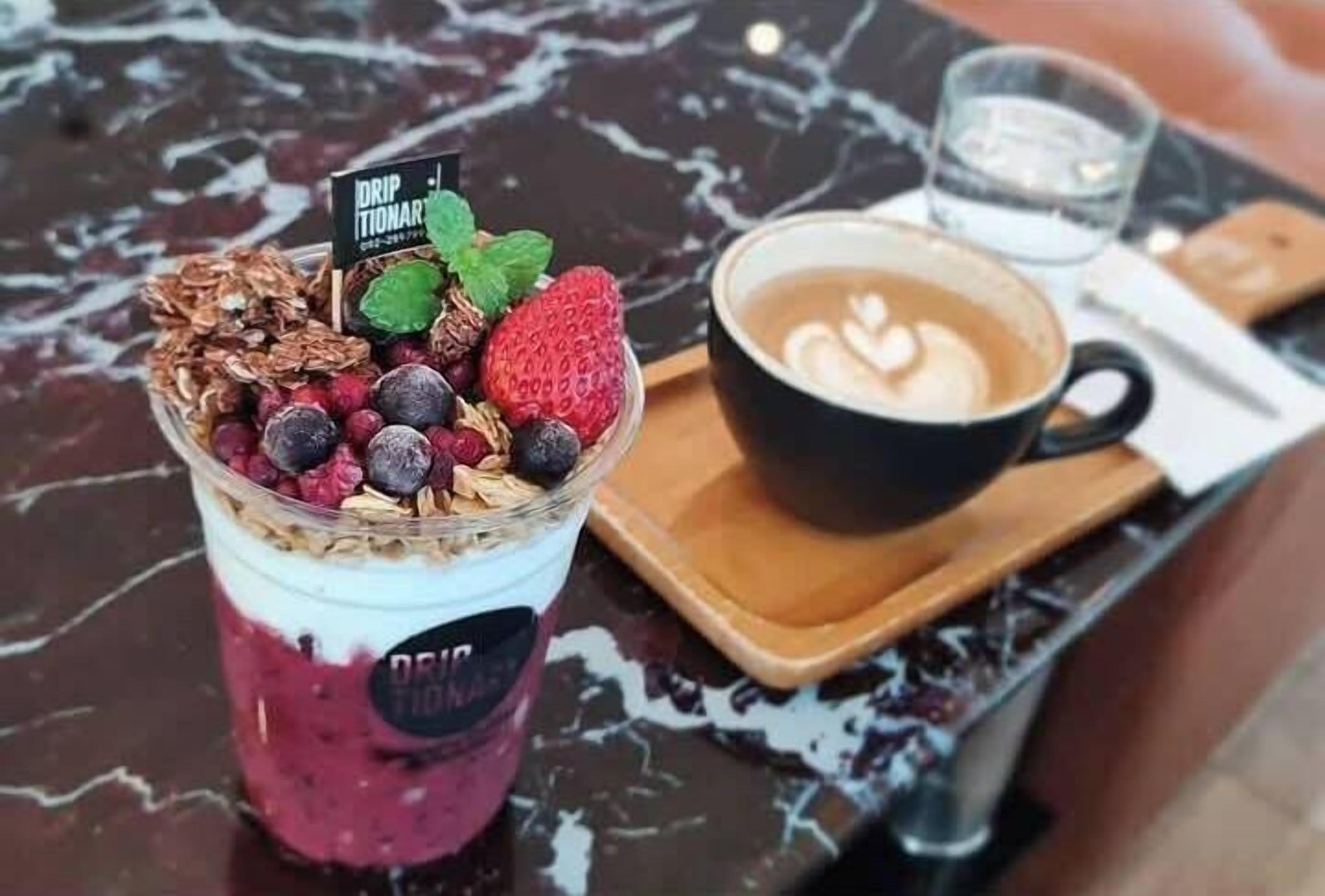 A dessert drink with berries and granola, a cup of coffee with latte art, and a glass of water on a marble table.
