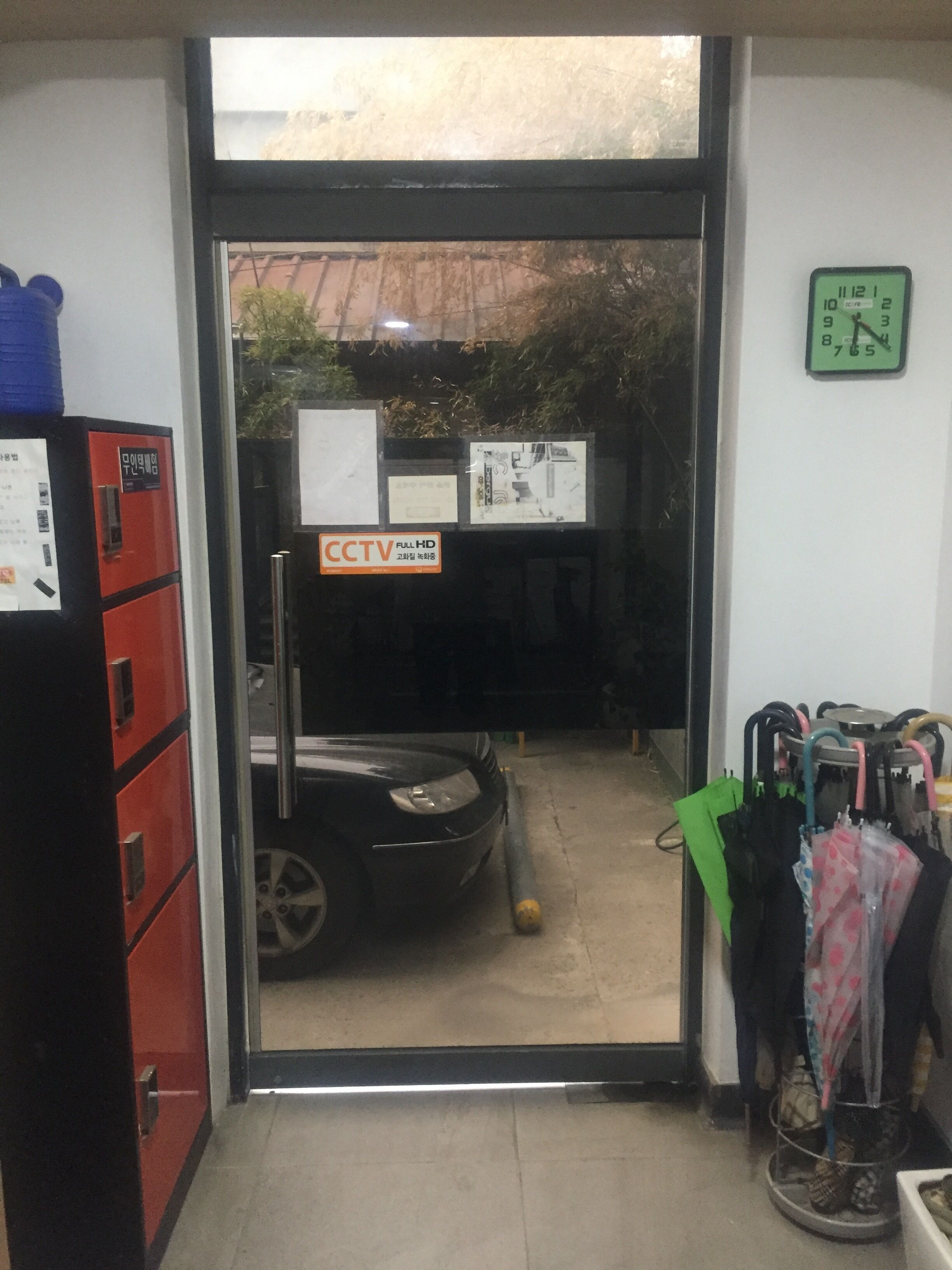 A doorway leading to a parking area with a CCTV sign, a clock on the wall, and a rack of umbrellas.