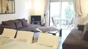 A living room with a brown sofa, a dining table with chairs, a flat-screen TV, and a balcony with a table and chairs.