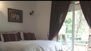 A bedroom with a bed, a painting on the wall, and a view of the outdoors through a window with curtains.