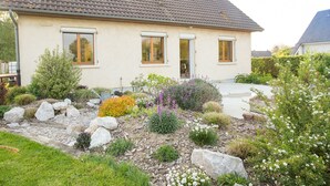 Ein zweistöckiges Haus mit Ziegeldach, einer Veranda und einem angelegten Garten mit verschiedenen Pflanzen und Steinen.