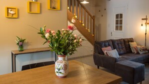 Ein Wohnzimmer mit einem Holztisch, einer Couch und einem Blumenstrauß. An der Wand sind gelbe Bilderrahmen und im Hintergrund eine Treppe zu sehen.