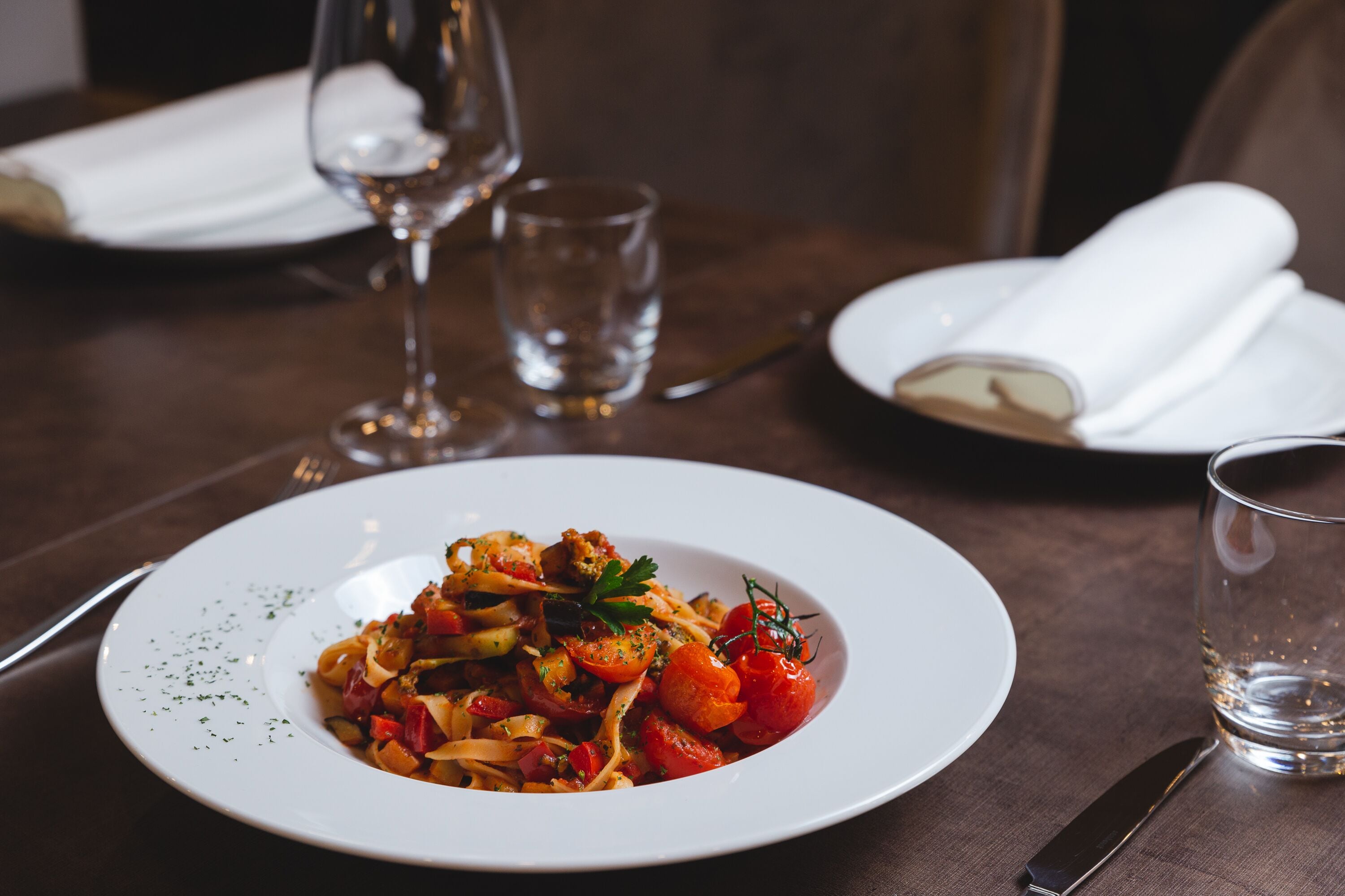 A dining table with a white plate of pasta, a glass of water, and a wine glass.