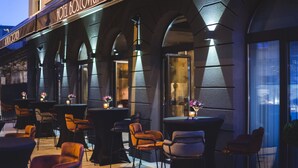 An outdoor seating area with tables and chairs, a sign reading "HOTEL BOSCOVICH," and a view of the cityscape through large windows. - Boscovich Boutique Hotel (Podgorica)