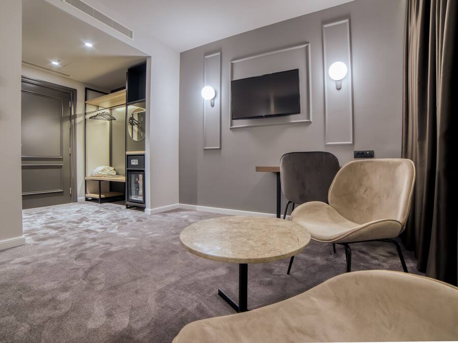 Quarto de hotel com uma mesa redonda, duas cadeiras, uma televisão fixada na parede e um espelho.