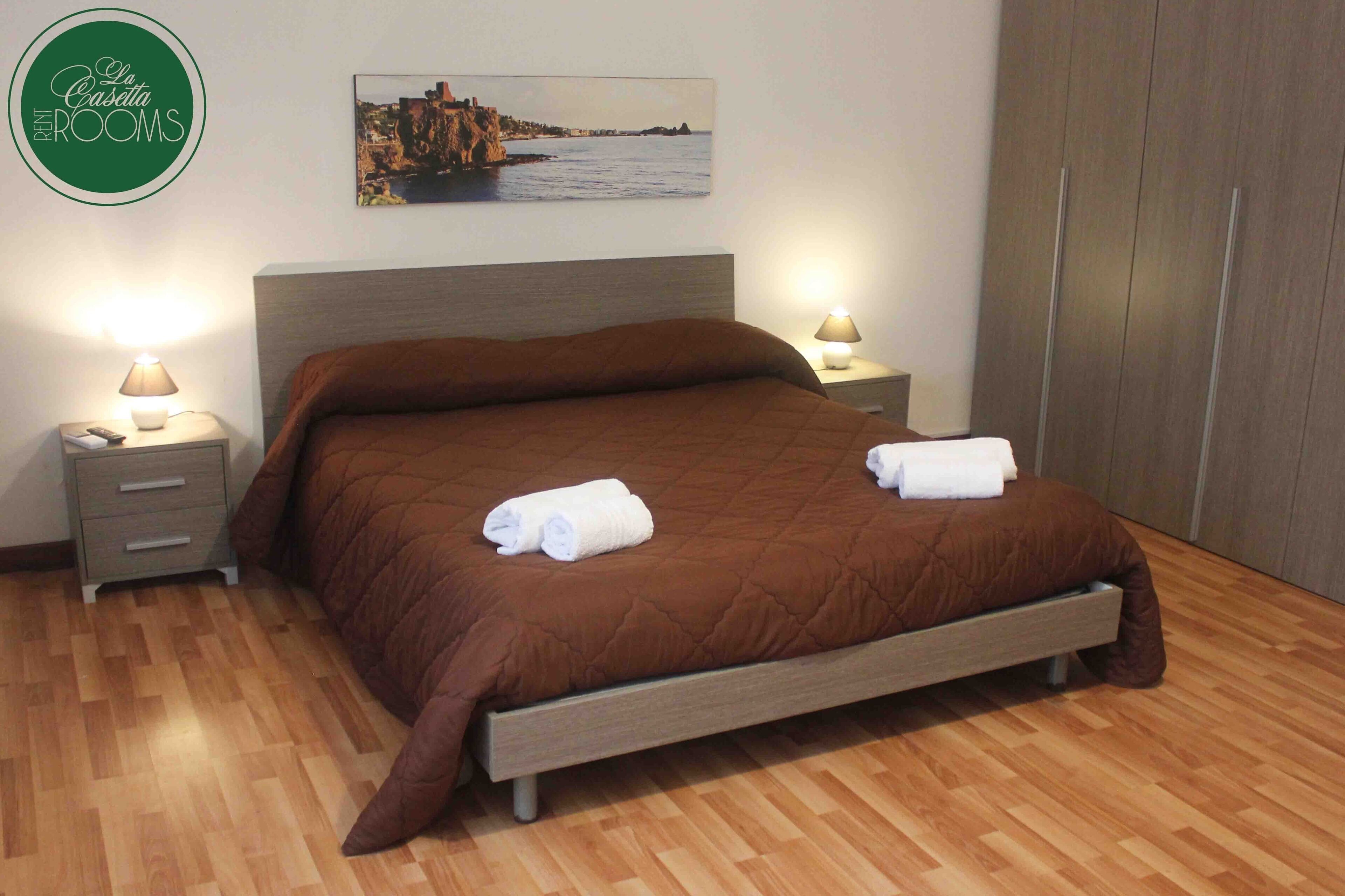 A neatly made bed with a brown quilt, two bedside tables with lamps, a wooden floor, and a wall-mounted picture of a coastal scene.
