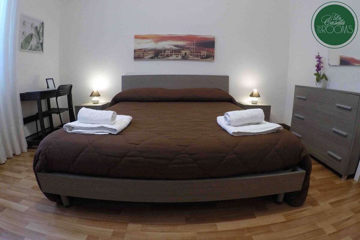 A neatly made double bed with a brown quilt, two bedside tables with lamps, a chair, a dresser, and a cityscape painting on the wall.