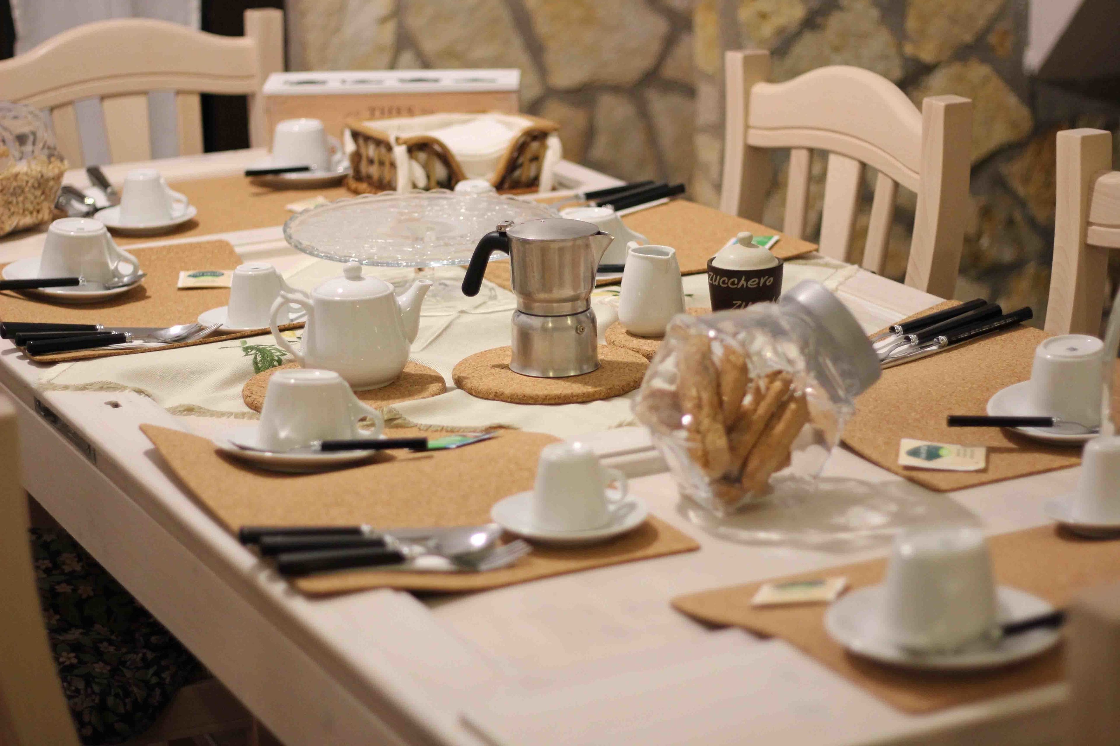 A dining table set with white cups, saucers, cutlery, and a coffee pot.