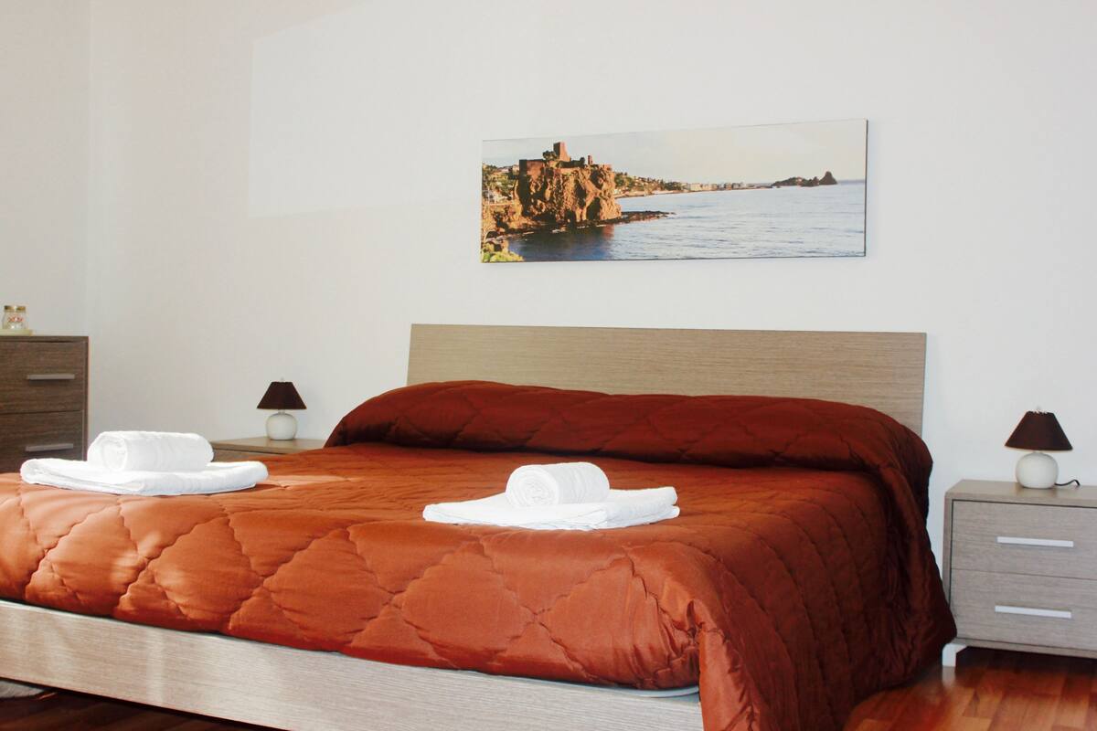 A neatly made bed with a red quilt, two white towels, and a painting of a coastal scene above it.
