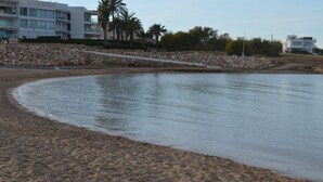 Ein Sandstrand mit ruhiger See, einer Steinmauer und modernen Gebäuden im Hintergrund.