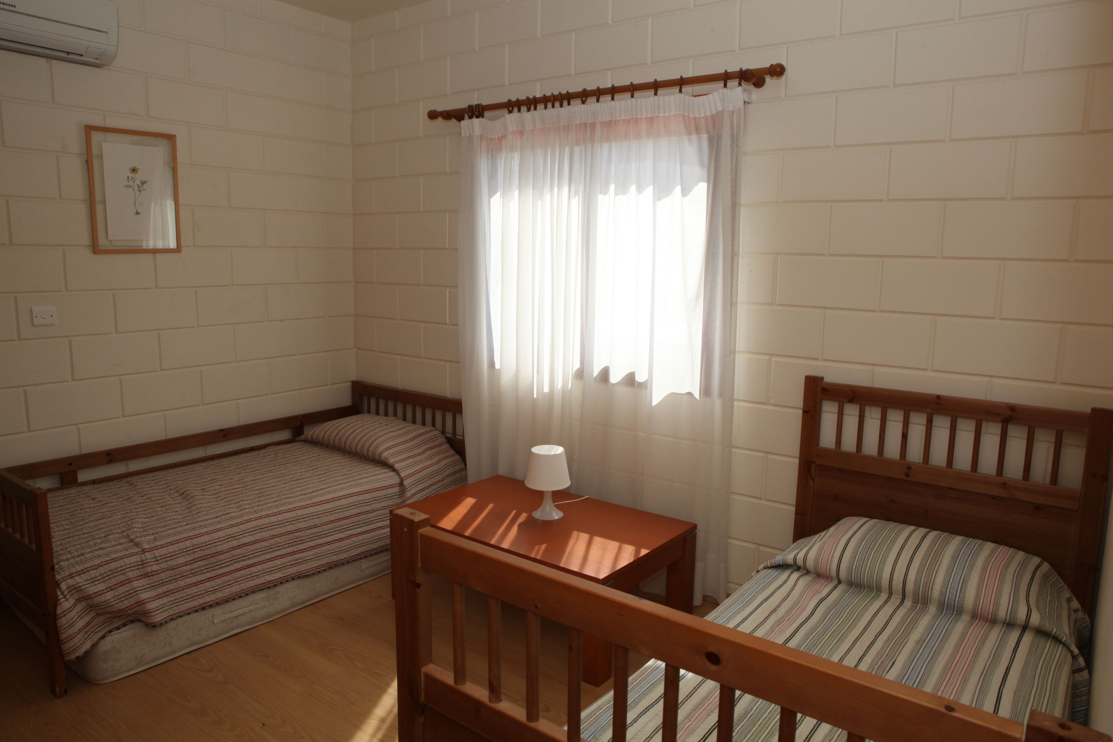 A room with two wooden beds, a small table with a lamp, a framed picture on the wall, and a window with curtains.