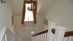 A staircase with white railings and a wooden handrail, leading to a room with a radiator, a window with brown curtains, and a chandelier.