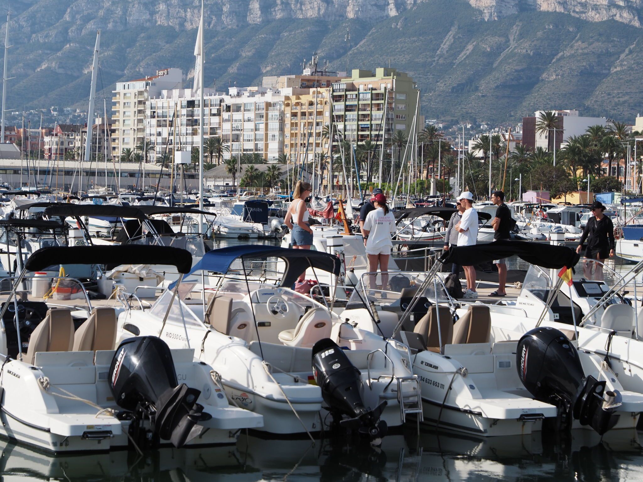 A marina with boats docked, a mountainous backdrop, and a building with balconies.