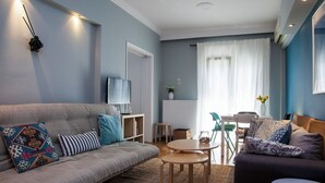 A modern living room with a grey sofa, a round coffee table, and a dark sofa with patterned cushions. A TV is mounted on the wall, and there is a dining area with a table and chairs. - Sunny & Happy Apartment Historic Center (Thessaloniki)