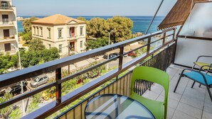 Een balkon met een glazen tafel en groene stoelen, uitkijkend op een straat met auto's en gebouwen, en een uitzicht op de zee.