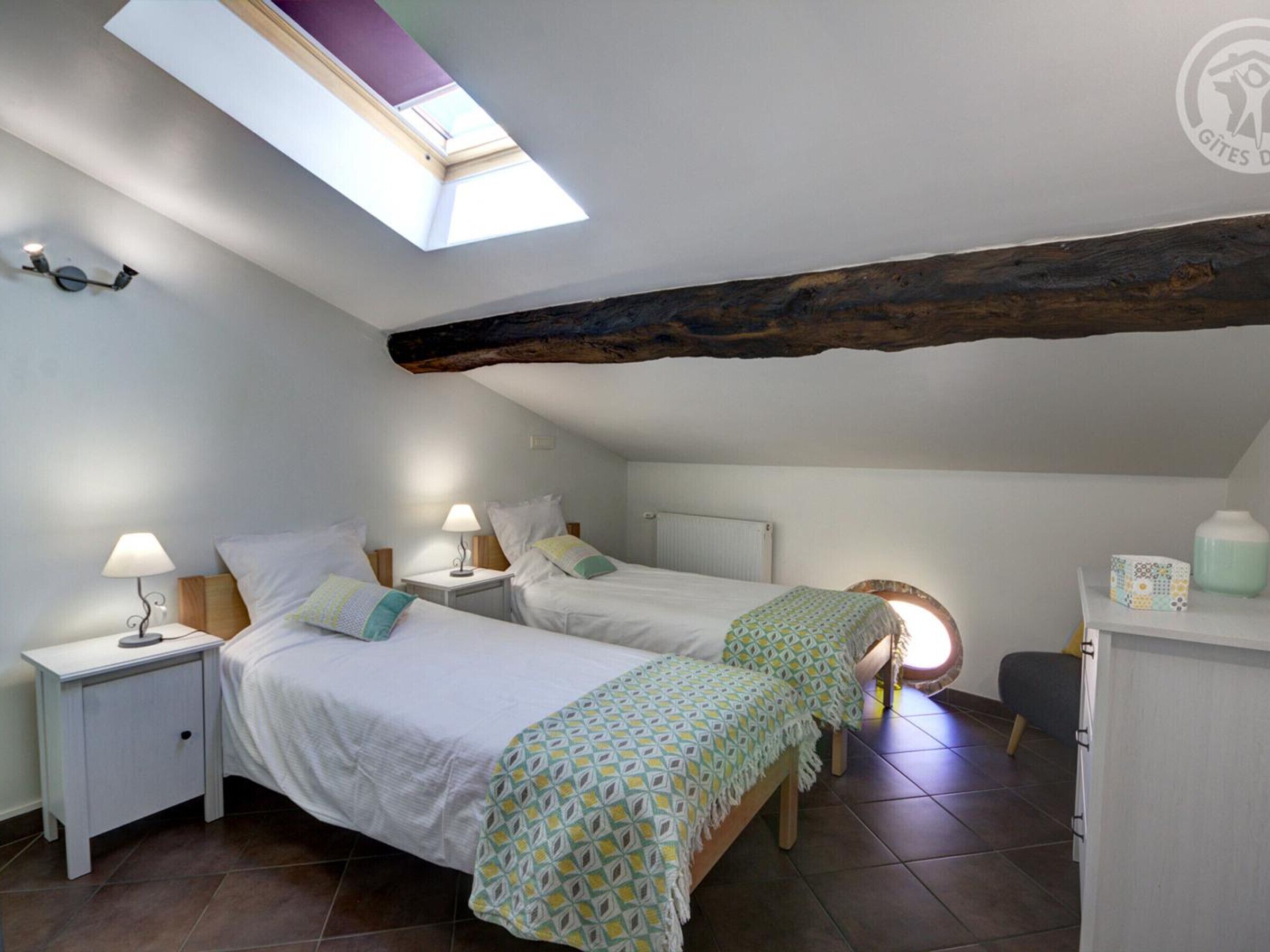 Une chambre mansardée chaleureuse, dotée de deux lits, d’une lucarne et d’un plafond avec une poutre en bois rustique.