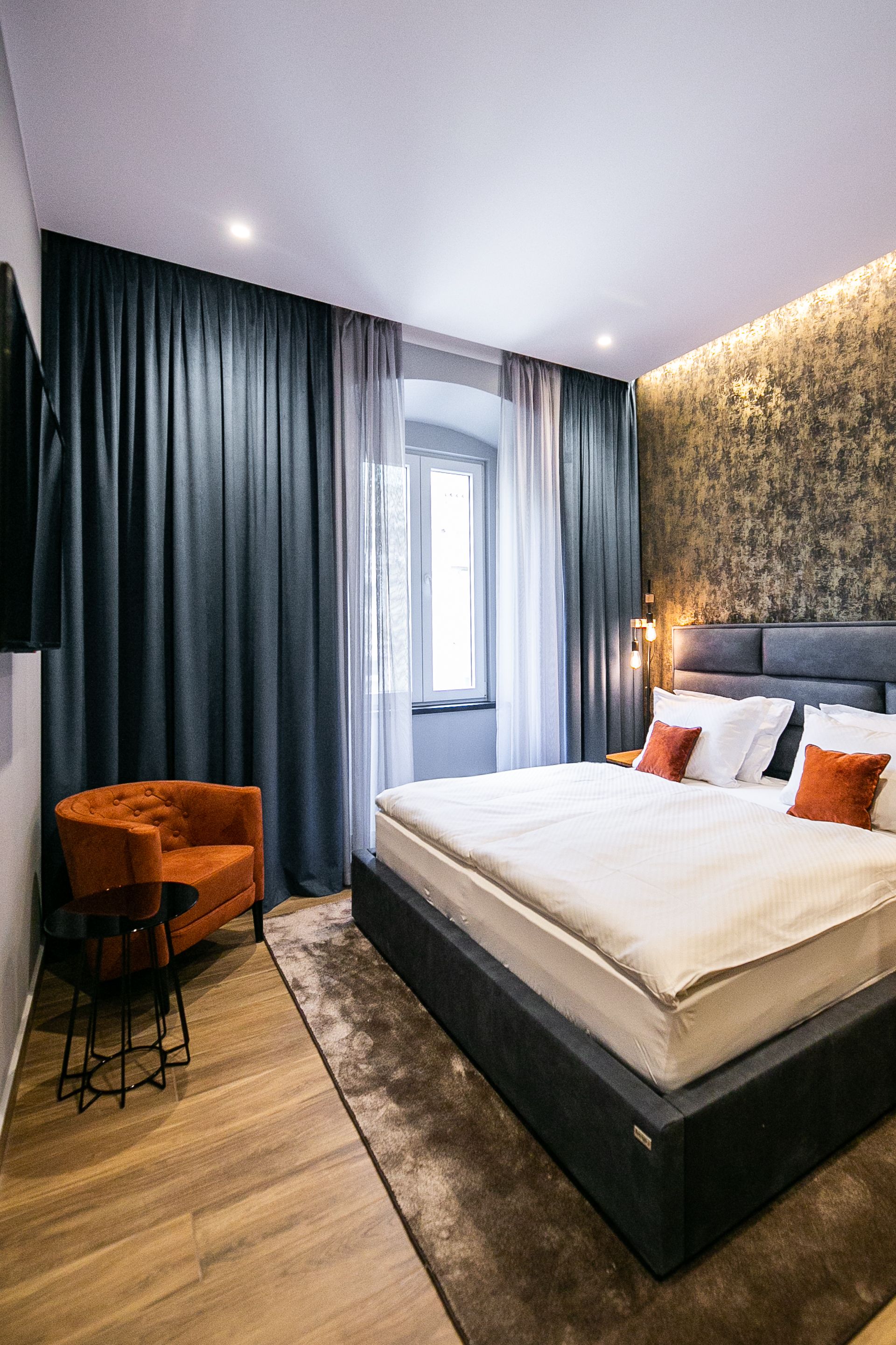 A modern hotel room with a large bed, a seating area with a chair, and a view of the outdoors through a window.