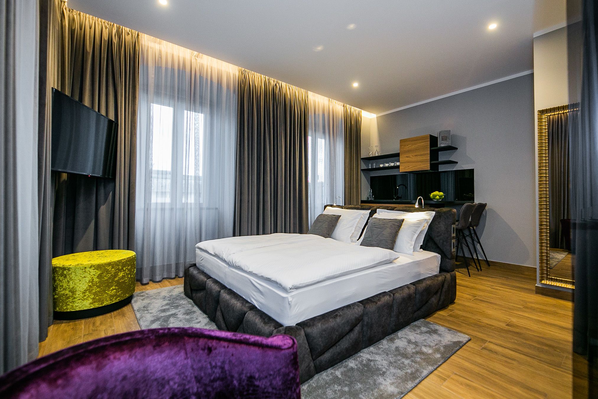 Een moderne slaapkamer met een groot bed, een flatscreen televisie, een kleine keukenhoek en een zithoek met een paarse stoel.