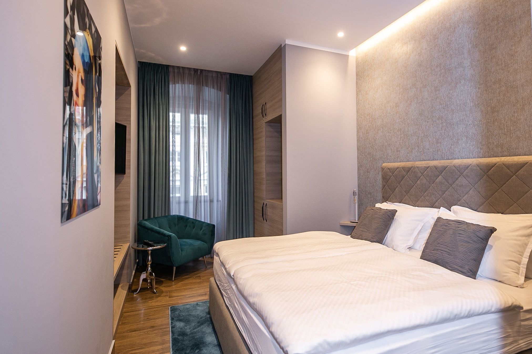 Una habitación de hotel moderna con una cama grande, un sillón verde, una mesita auxiliar pequeña y un ventanal con cortinas.
